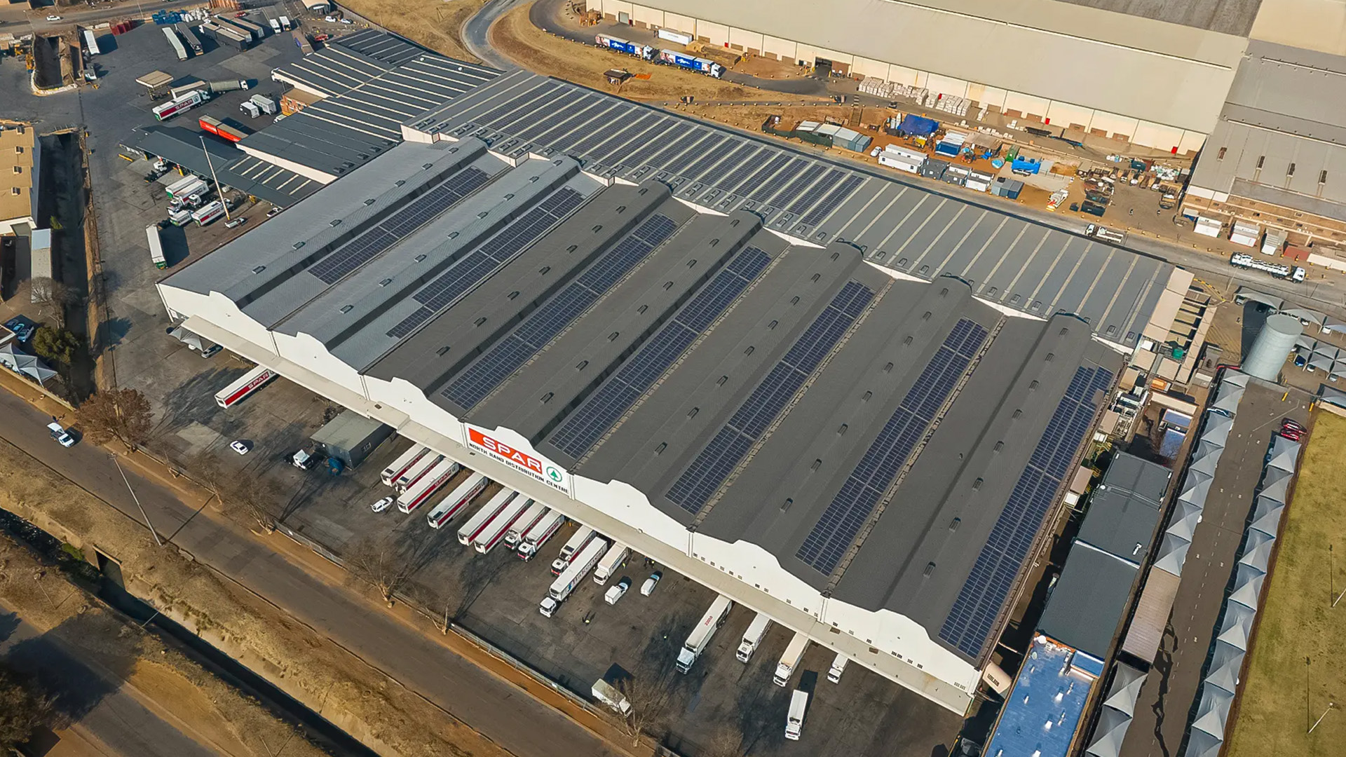 Aerial view of a large SPAR distribution center, showcasing its expansive rooftop solar panels and busy truck loading docks.