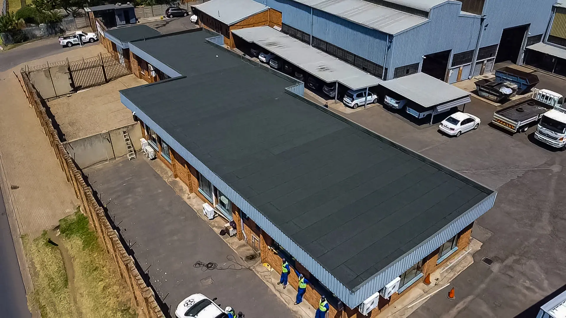 Detailed aerial view of a large industrial building with a newly installed dark gray industrial flat roof, cars, and surrounding structures.
