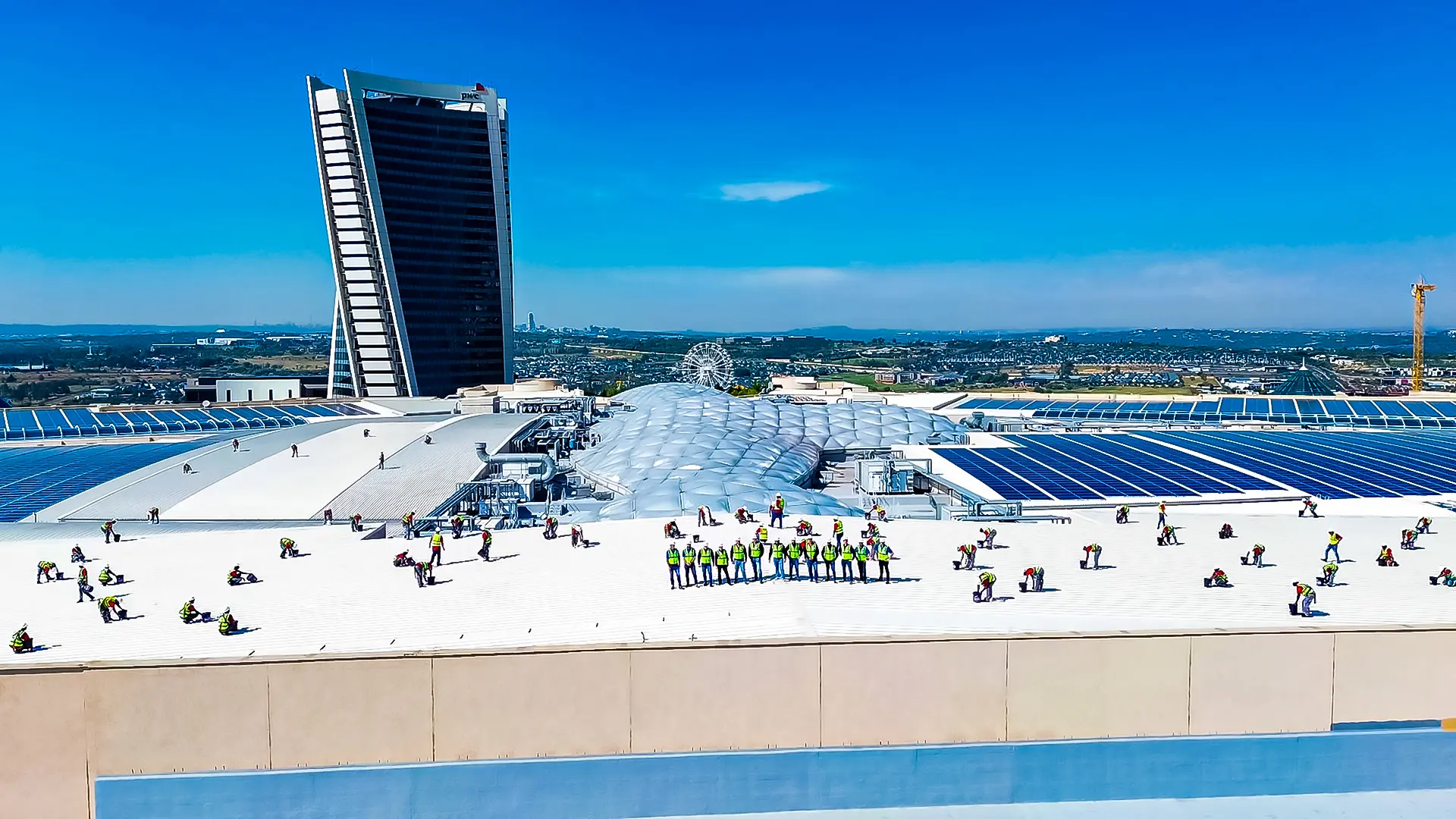 Large crew installs shopping mall waterproofing on expansive white rooftop, solar panels, and PwC tower in city.