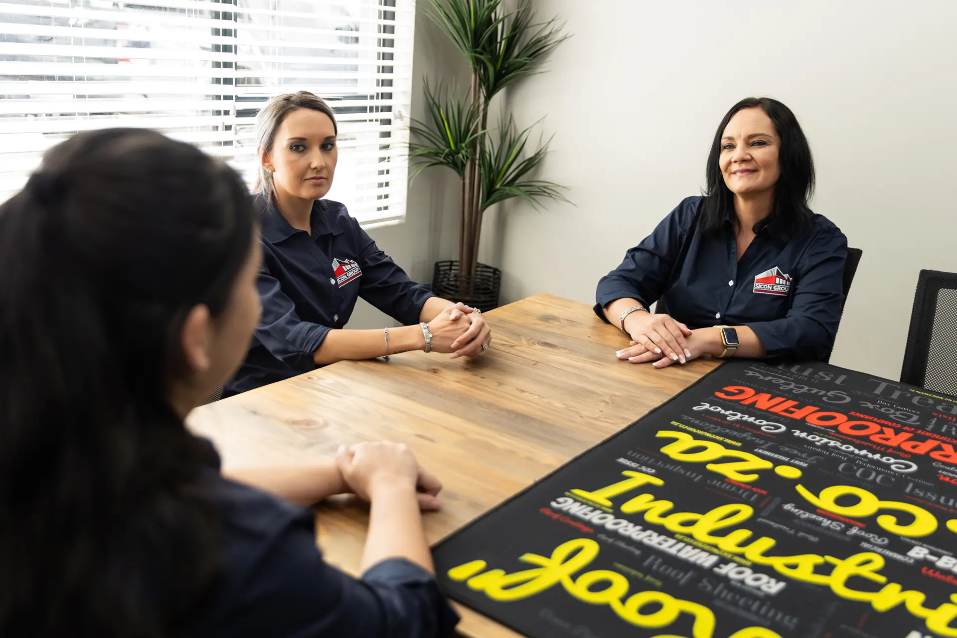 Two professional women from Sicon Group in branded shirts discussing business at a wooden table, representing a leading waterproofing company near you.
