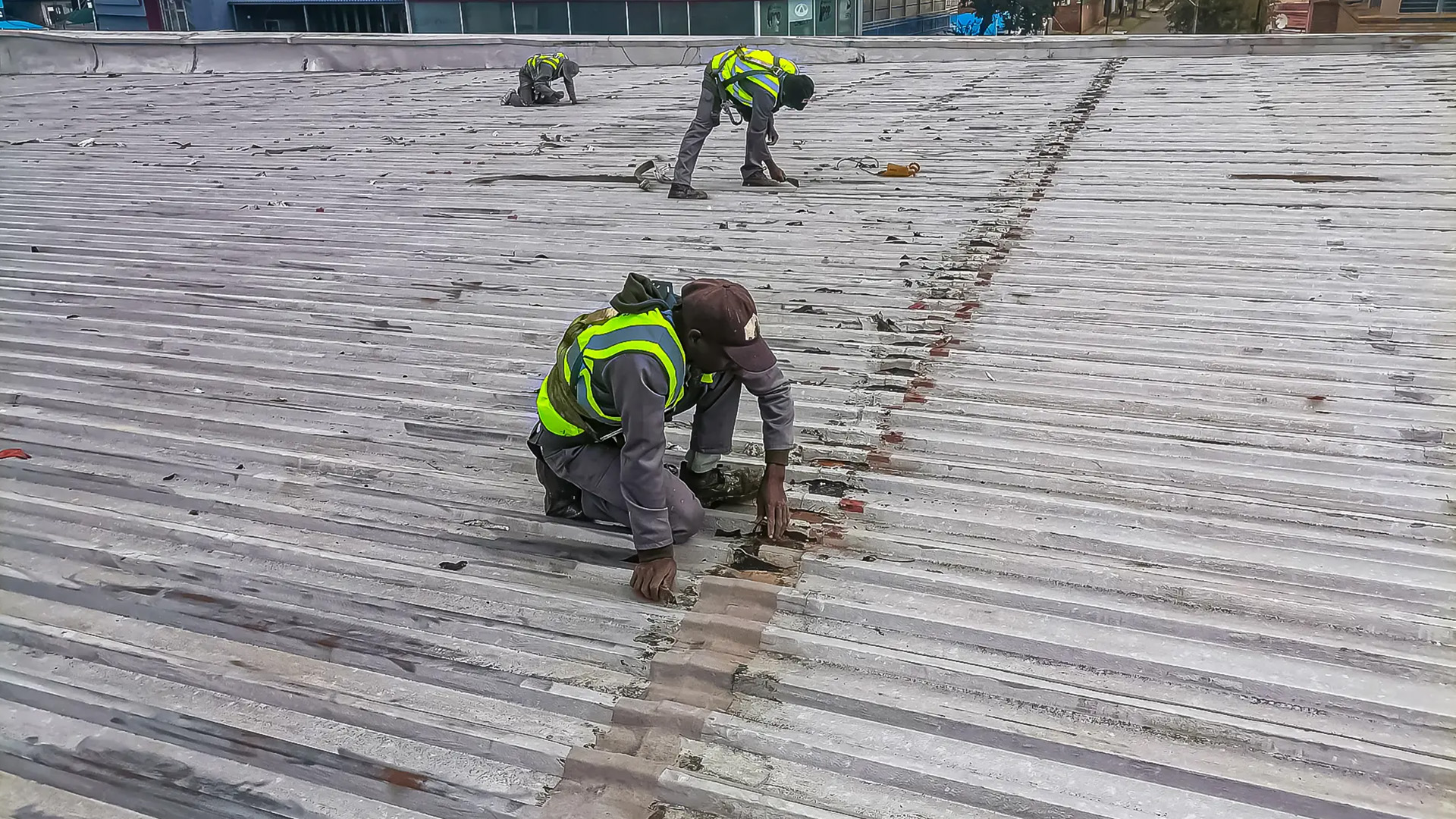 Skilled roofers in safety vests perform corrugated metal roof repair on a commercial building under a cloudy sky.