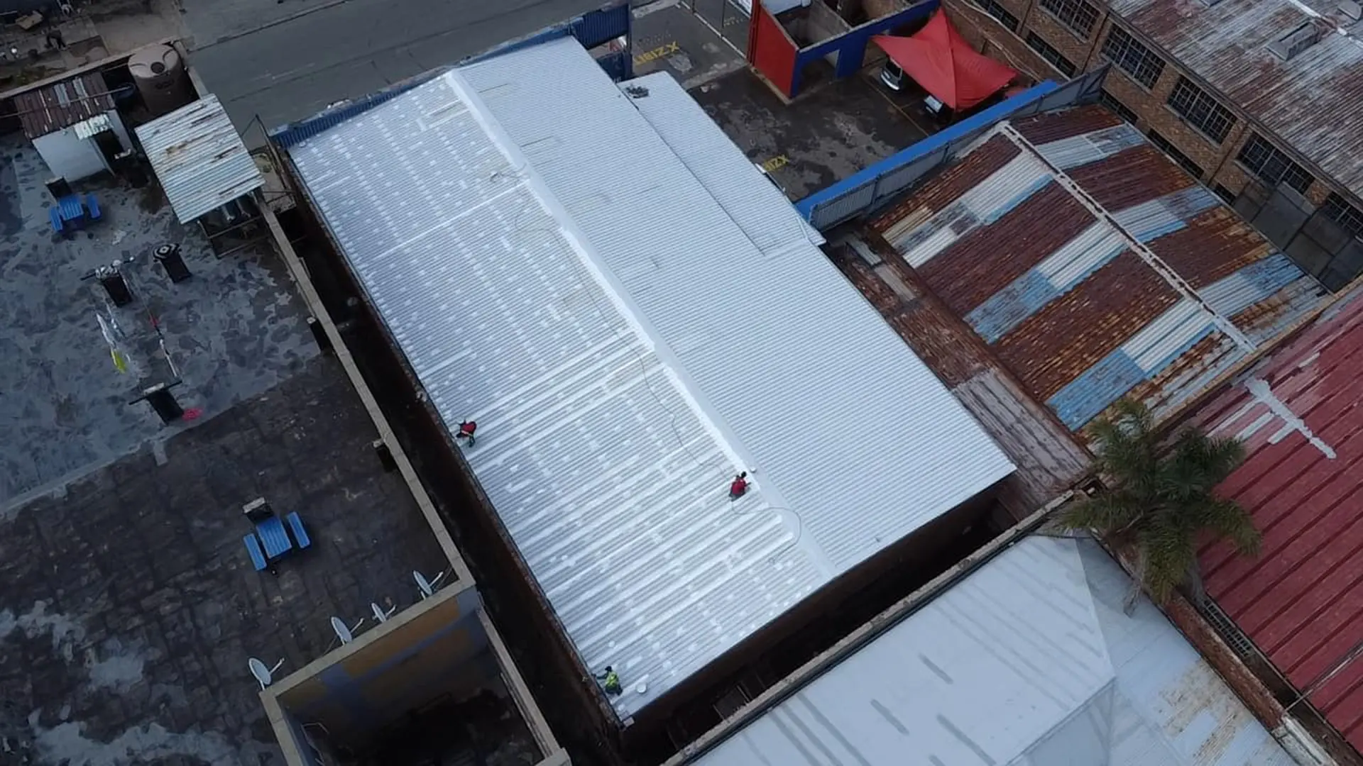 Aerial view of workers applying specialized roofing waterproofing to a large corrugated metal industrial building roof.