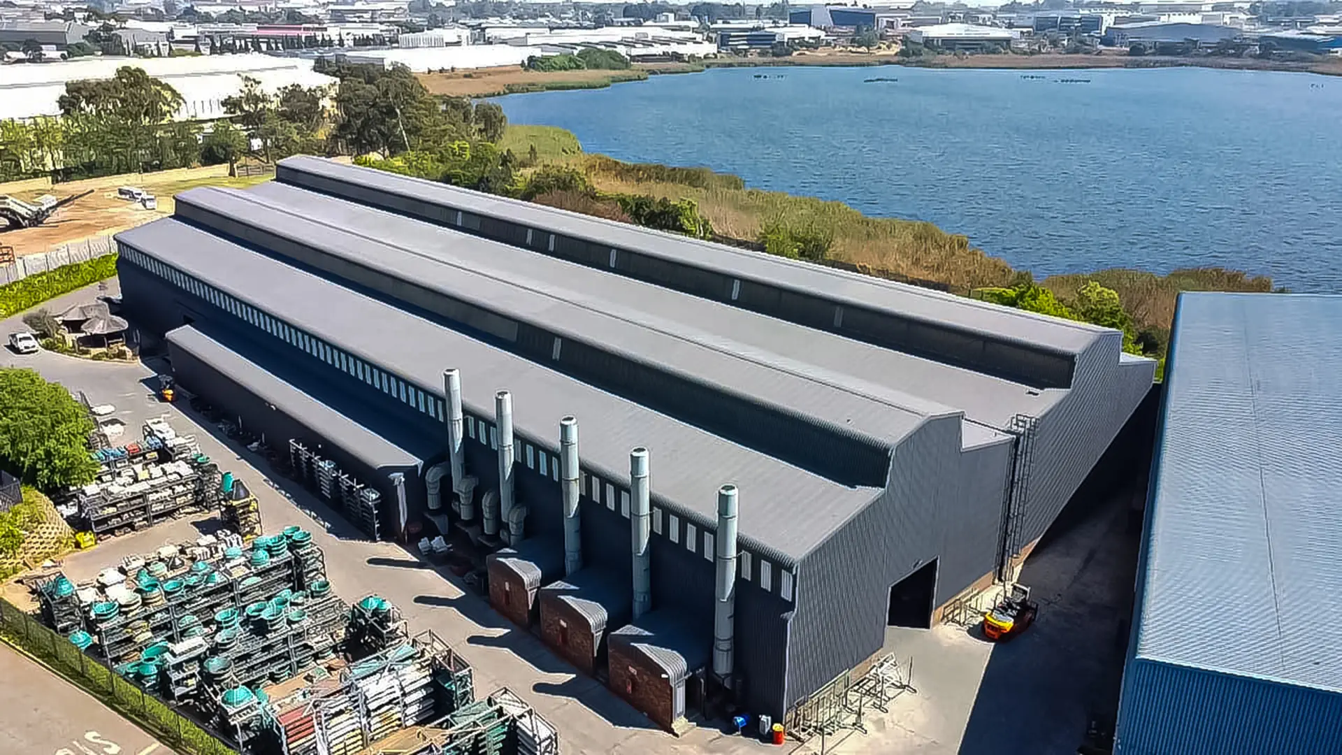 Large industrial facility with modern grey metal roofing sheets by a lake, showcasing effective waterproofing for industrial roofs.